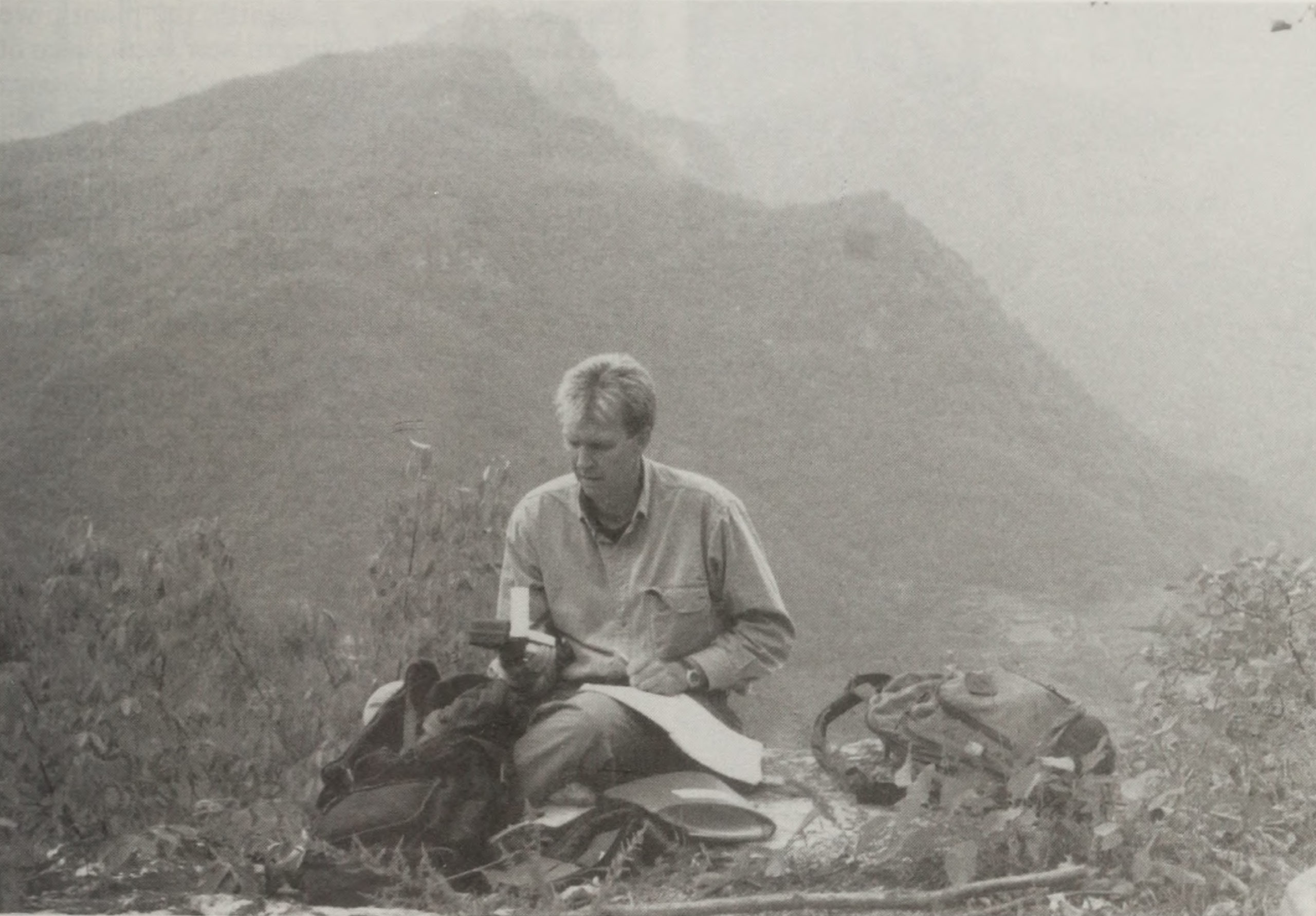 Black-and-white photograph of man sitting on ground with supplies arrayed around him