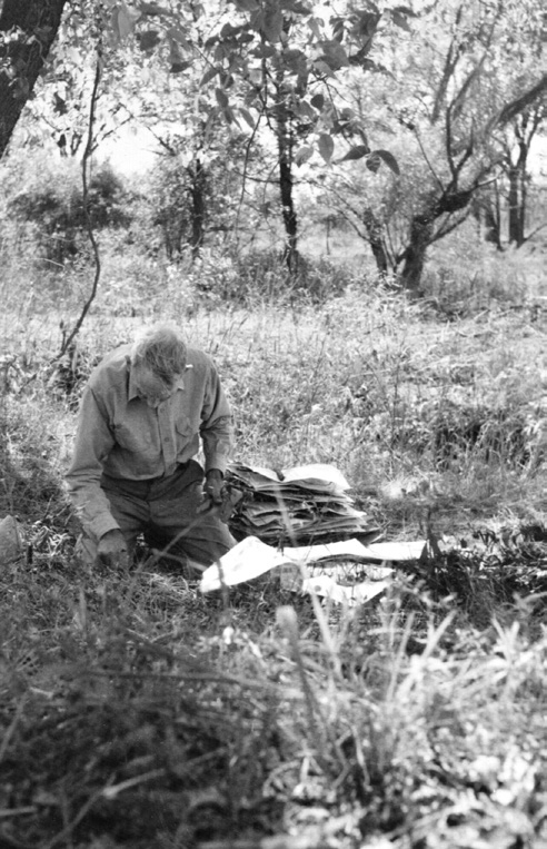 Black-and-white photograph of man kneeling on ground with herbarium specimen