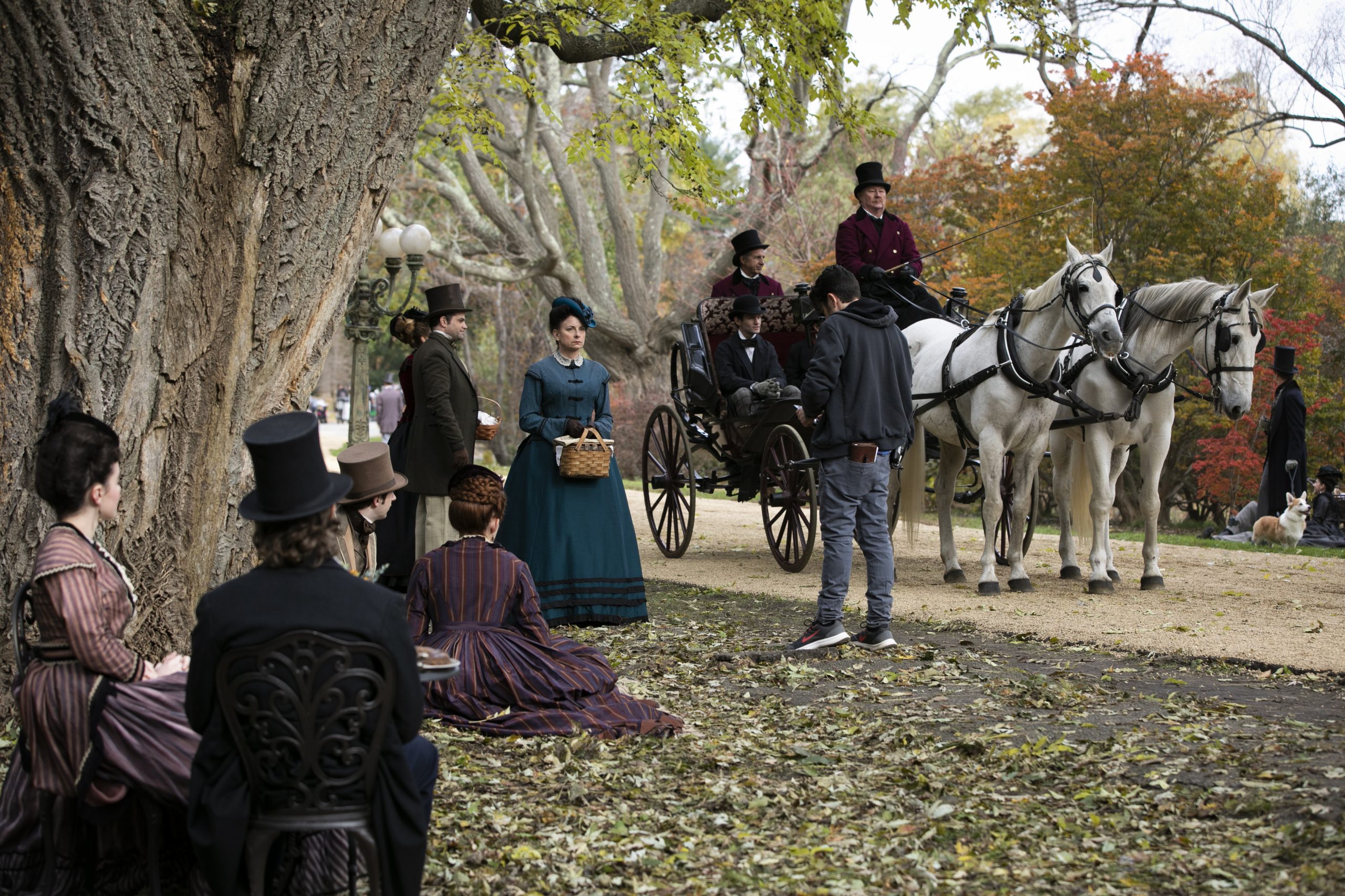 Actors and actresses in the Arnold Arboretum for the filming of Little Women. Half a dozen people in fashion of the times stand and sit around a tree with a horse-drawn carriage in the background.