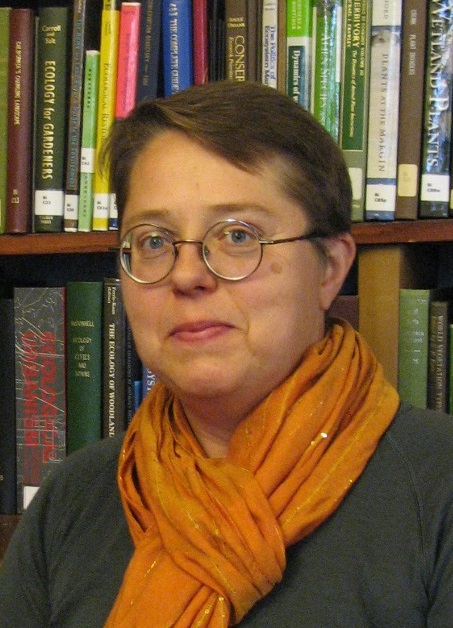 Photograph of a person wearing an orange scarf stand in front of a bookcase.
