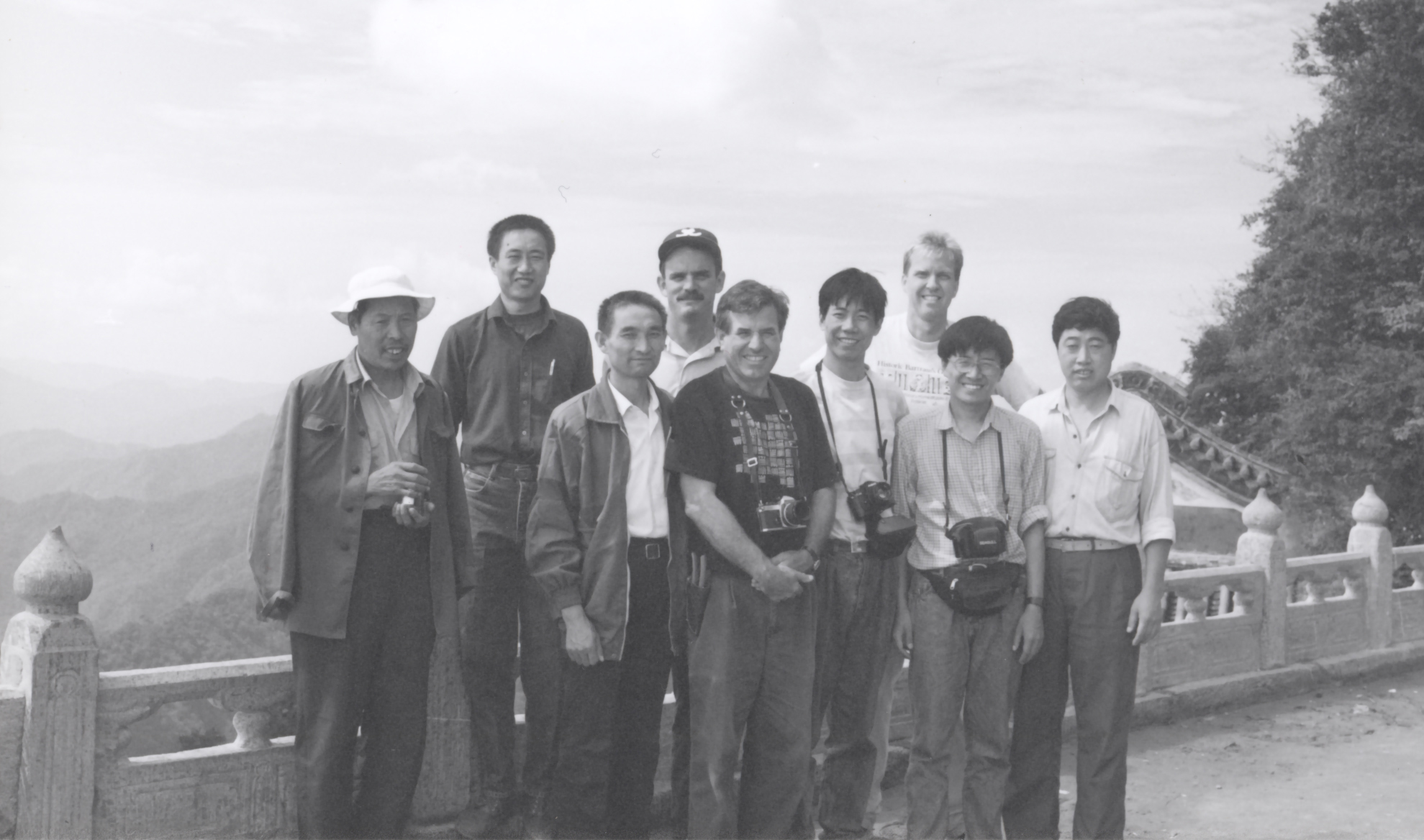 Nine men posed at a mountain overlook, several wear cameras around their necks.