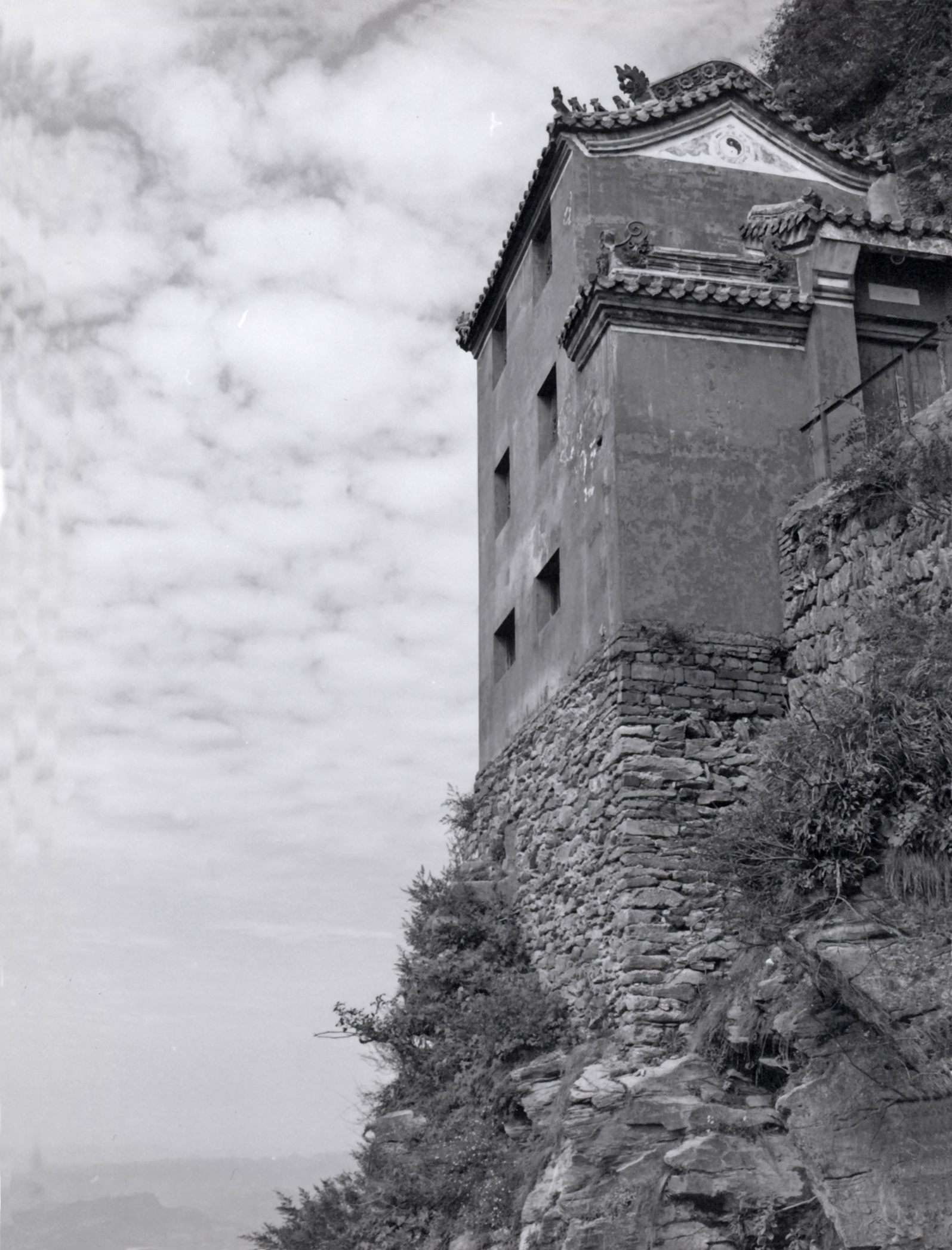 A light colored stone building is built onto a cliff face. The sky with clouds is seen beyond in this black and white photograph.