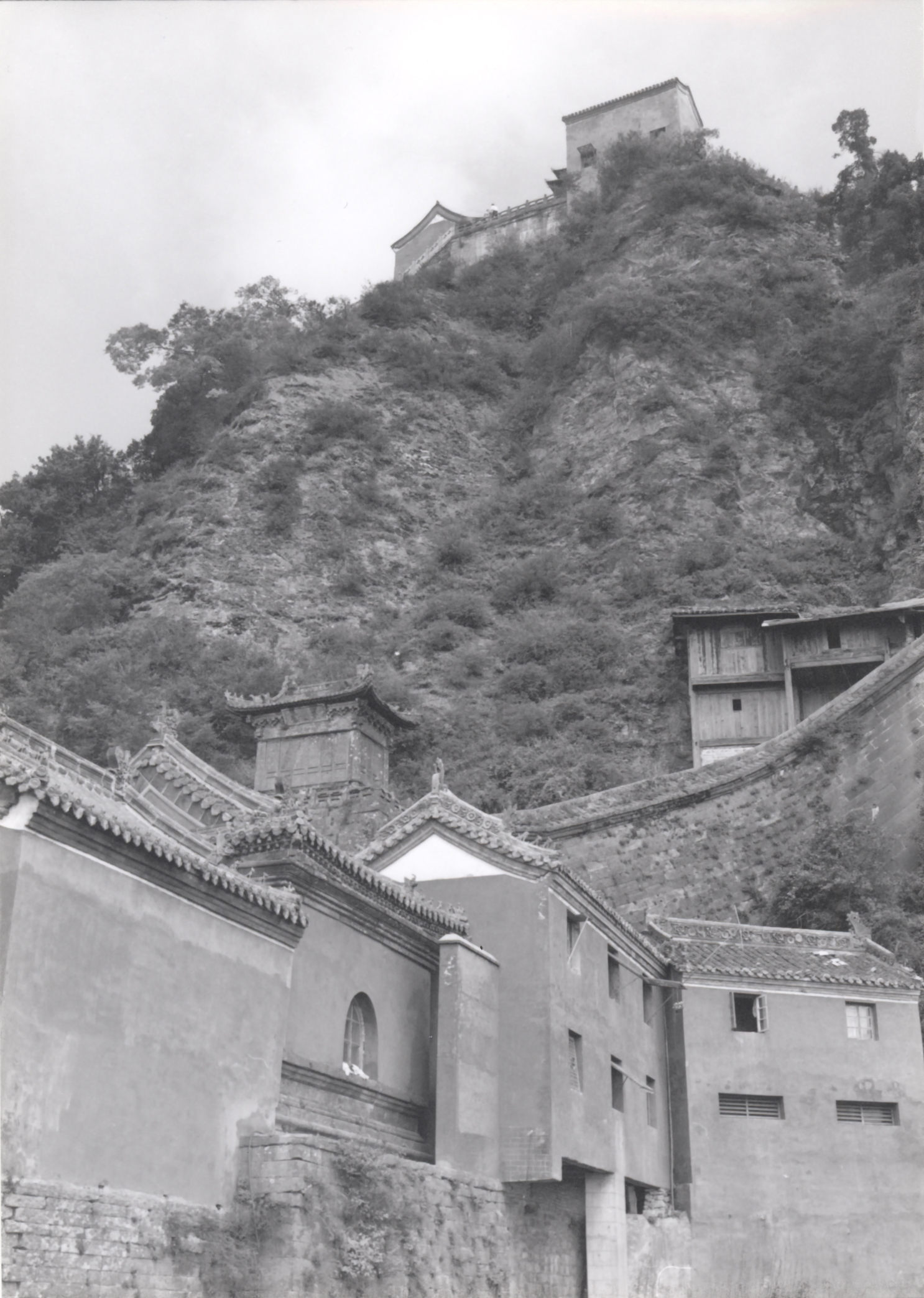 Stone buildings with tile roofs at the foot of a vegetation covered cliff with a building at the top of the cliff and sky beyond.