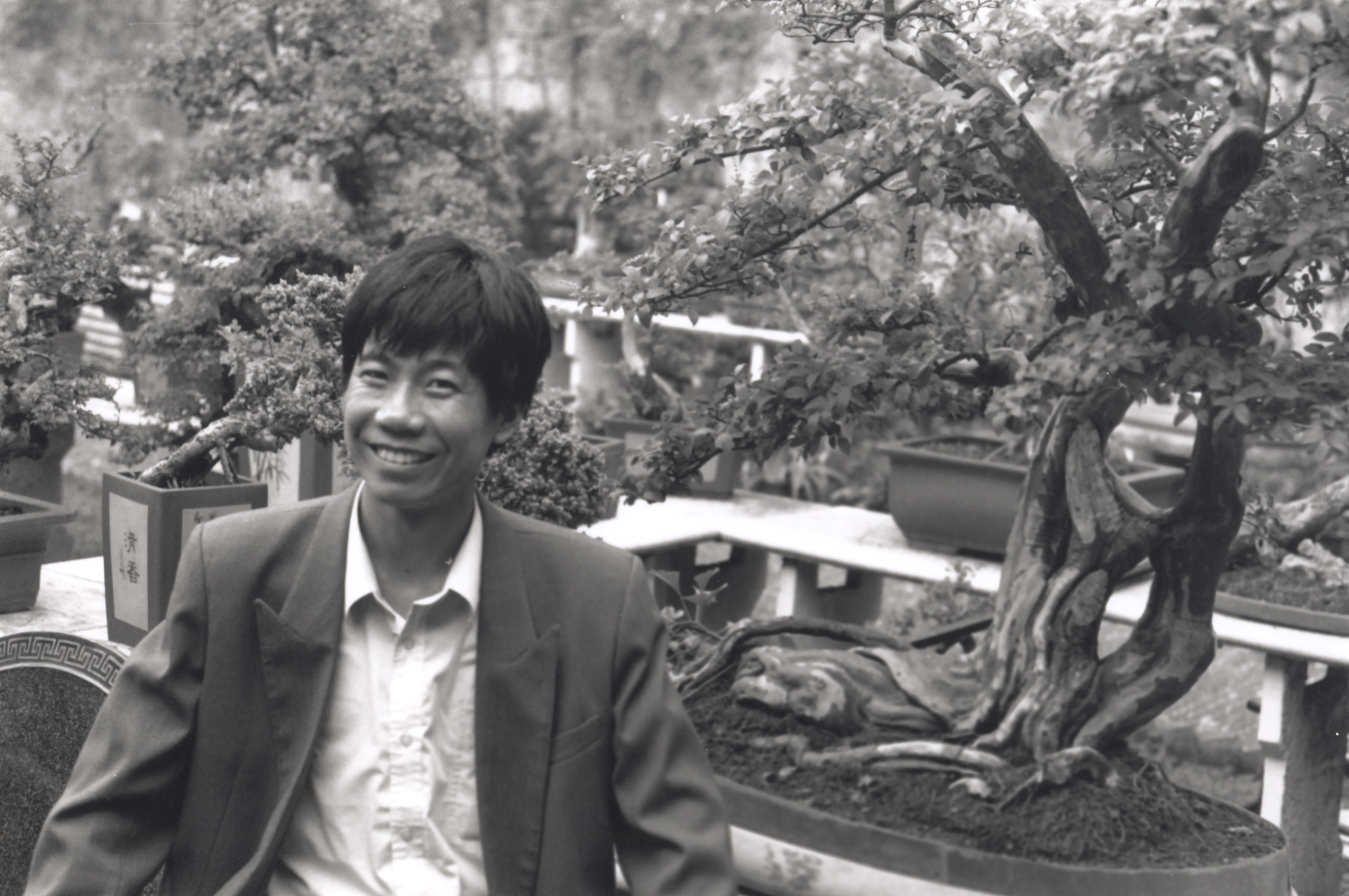 A man, viewed from the waist up, wearing a white buttoned down shirt and grey sports jacket, stands in front of miniature potted trees on a shelf.