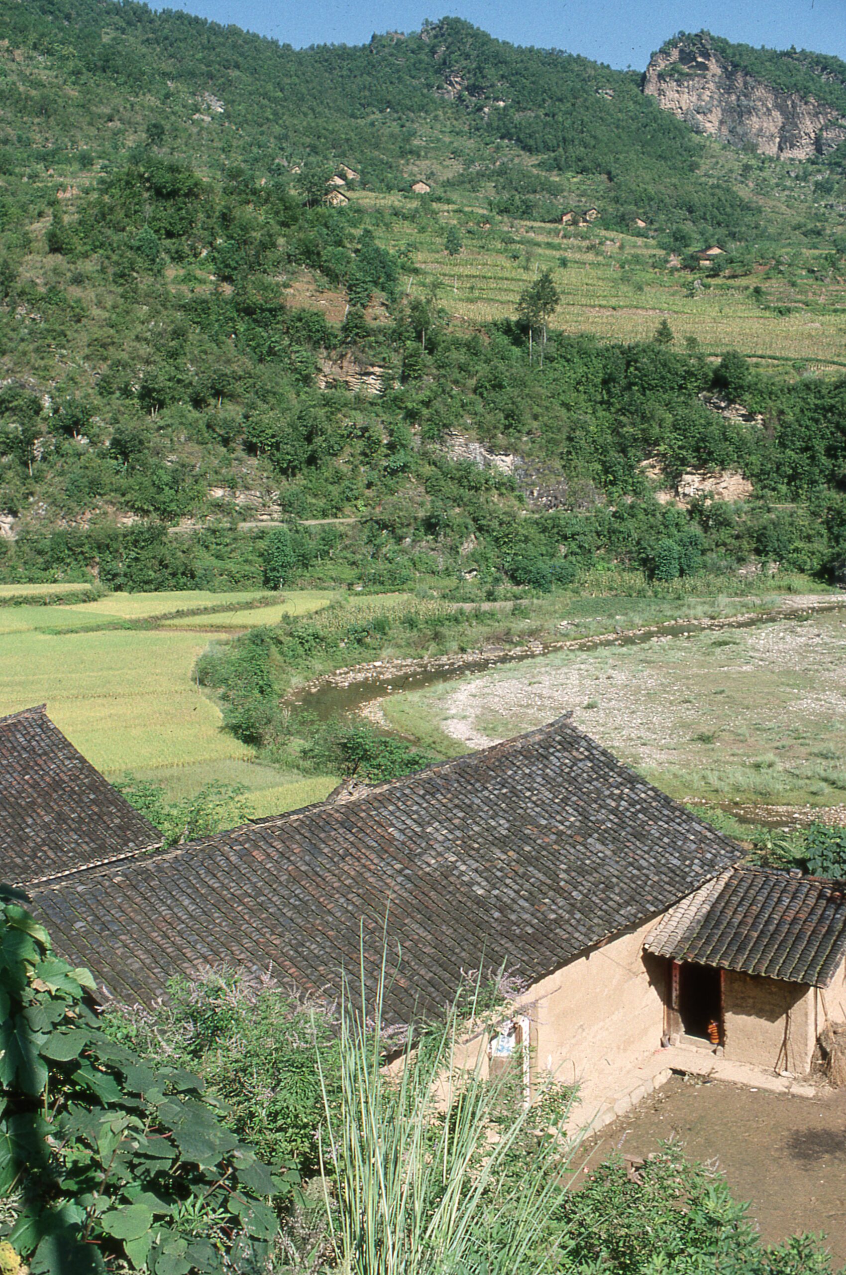 View of an agricultural area with a tan building with a brown roof in the foreground and terraced field on a mountainside beyond.